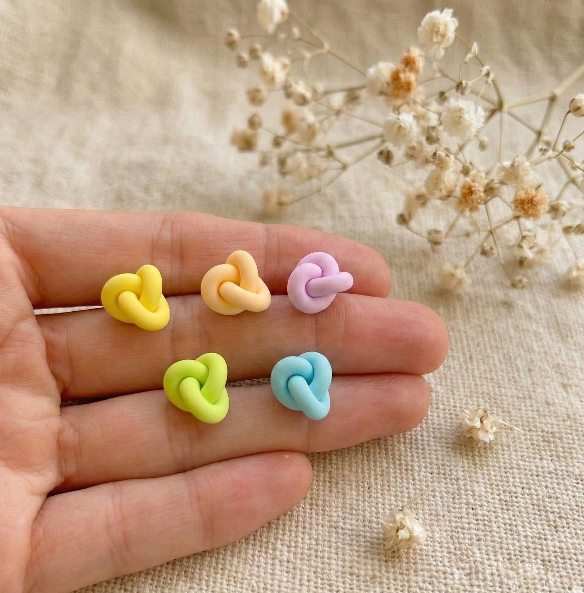Hand holding four pairs of small knot-shaped stud earrings in pastel yellow, pink, lilac and blue - minimalist handmade polymer clay earrings by Clay by KM